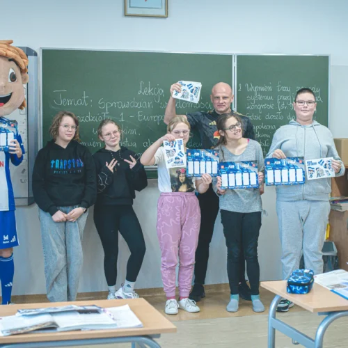 Na zdjęciu znajduje się grupa uczniów i nauczycieli w klasie, trzymających plakaty i sygnalizujących radość, wśród nich postać w kostiumie z dużą głową.