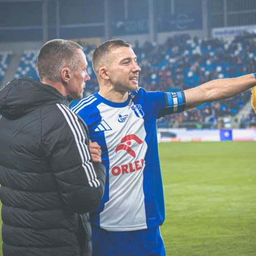 Na zdjęciu znajduje się piłkarz w mundurze drużyny sportowej, który rozmawia z drugim mężczyzną, trzymając flagę klubową na stadionie.