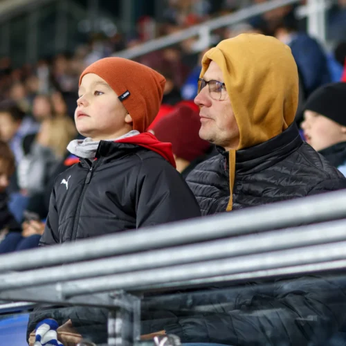 Na zdjęciu widać chłopca w pomarańczowej czapce oraz mężczyznę w żółtej bluzie, obydwaj z uwagą obserwują wydarzenie na boisku, otoczeni tłumem innych widzów.