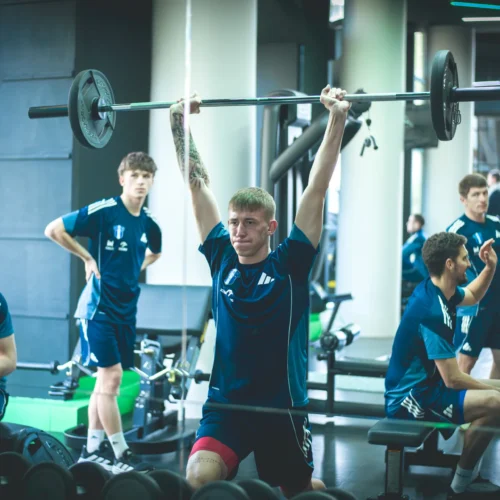 Na zdjęciu znajduje się grupa młodych sportowców w treningu, z jednym z nich wykonującym ćwiczenie siłowe z ciężarem nad głową w nowoczesnej siłowni.