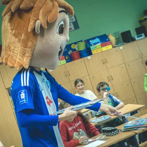 Na zdjęciu przedstawiono osobę w przebraniu postaci z dużą głową, rozdającą książki w klasie pełnej dzieci.