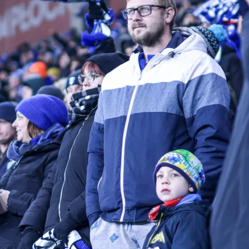 Na zdjęciu widoczna jest grupa kibiców, w tym mężczyzna i dziecko, którzy z zaangażowaniem obserwują wydarzenie sportowe na trybunach.