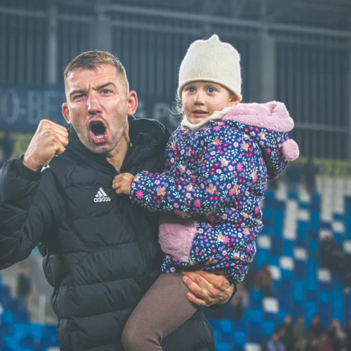 Na zdjęciu widać mężczyznę z radością unoszącego na rękach małą dziewczynkę, oboje cieszą się z emocjonującego momentu na stadionie.