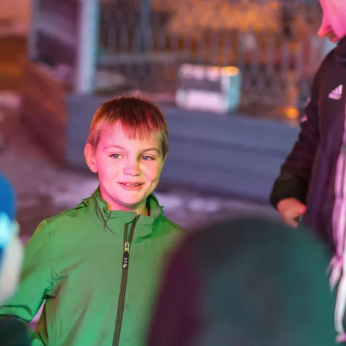 Na zdjęciu widać uśmiechniętego chłopca w zielonej kurtce, rozmawiającego z innymi dziećmi w otoczeniu kolorowych świateł.