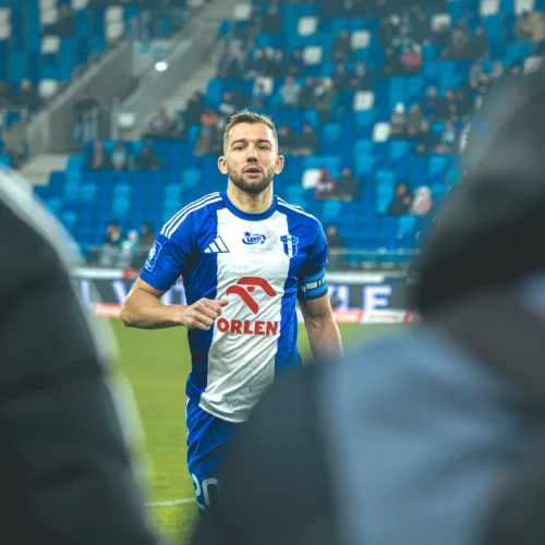 Na zdjęciu widoczny jest piłkarz w niebieskiej koszulce, który podchodzi do kamery na stadionie podczas meczu.