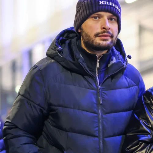 Mężczyzna w ciemnej kurtce puchowej i zimowej czapce stoi na ulicy w miejskim otoczeniu.