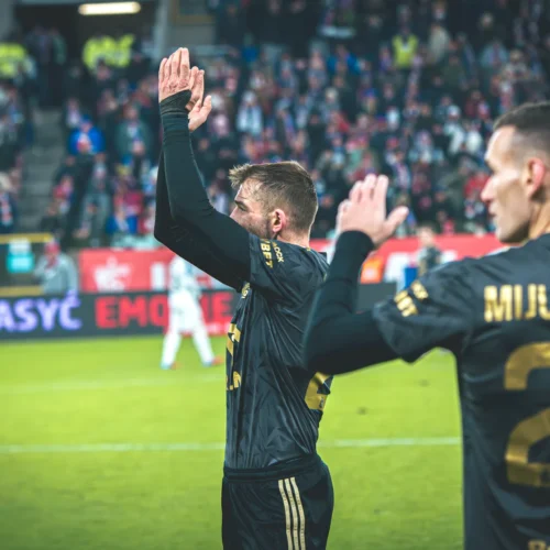 Na zdjęciu widać dwóch piłkarzy na boisku, którzy machają do kibiców w tle, podczas emocjonującego meczu.