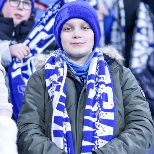 Na zdjęciu widoczny jest chłopiec ubrany w szalik i czapkę w kolorze niebieskim, stojący w tłumie kibiców na stadionie.