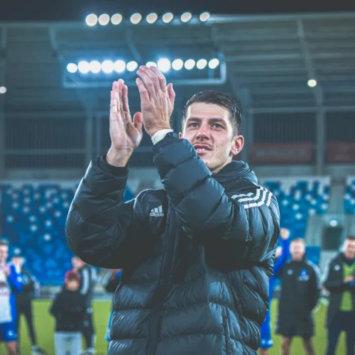 Mężczyzna w czarnej kurtce klaska w dłonie, stojąc na boisku, z radosnymi kolegami drużynowymi w tle.