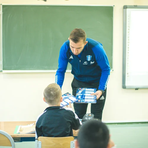 Na zdjęciu widać mężczyznę w niebieskiej kurtce, który wręcza książki uczniowi siedzącemu w klasie, podczas gdy na tablicy w tle widać zapisany czas.