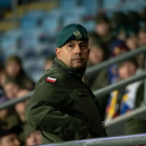 Mężczyzna w zielonej kurtce wojskowej z beretem i polską flagą, obserwujący wydarzenie z trybun.