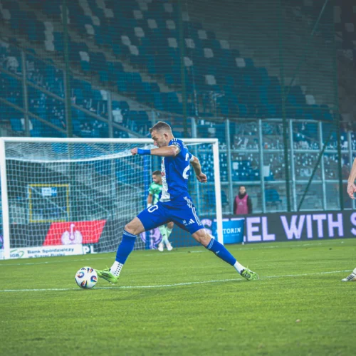 Na zdjęciu widać piłkarza w niebieskim stroju wykonującego strzał na bramkę, z zawodnikami przeciwnika w pomarańczowych strojach w tle.