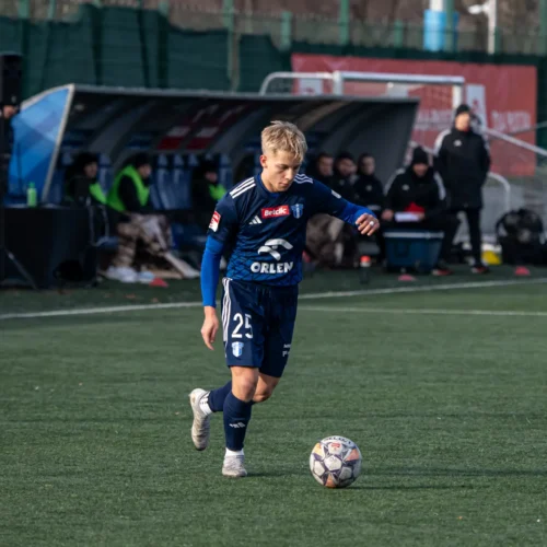 Młody piłkarz w niebieskim stroju z numerem 25 na koszulce zbliża się do piłki na boisku piłkarskim.