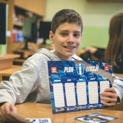 Chłopiec w szarym swetrze uśmiecha się, trzymając w rękach plan lekcji w klasie.