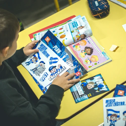 Na zdjęciu widać chłopca przeglądającego kolorowe książki i materiały edukacyjne na żółtym stole.