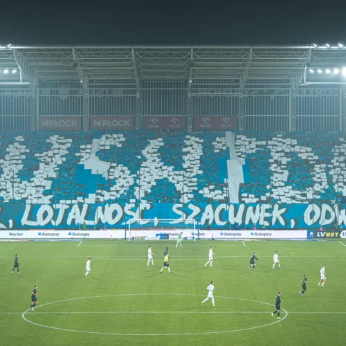 Na zdjęciu znajduje się stadion piłkarski, na którym kibice prezentują transparent z hasłem "HONOR, LOJALNOŚĆ, SZACUNEK, ODWAGA", podczas meczu drużyn grających na zielonej murawie.
