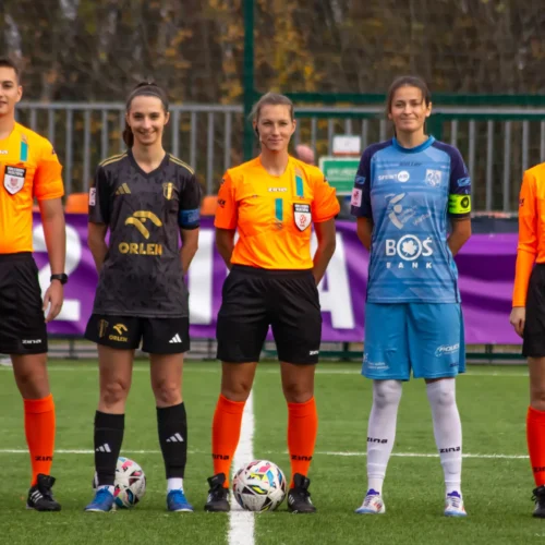 Na zdjęciu znajduje się grupa sędziów i zawodniczek środku boiska, przygotowujących się do meczu piłkarskiego.