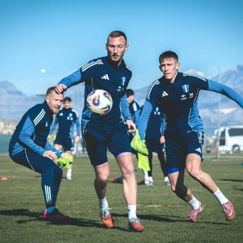 Grupa piłkarzy trenuje na boisku w słoneczny dzień, koncentrując się na grze z piłką.