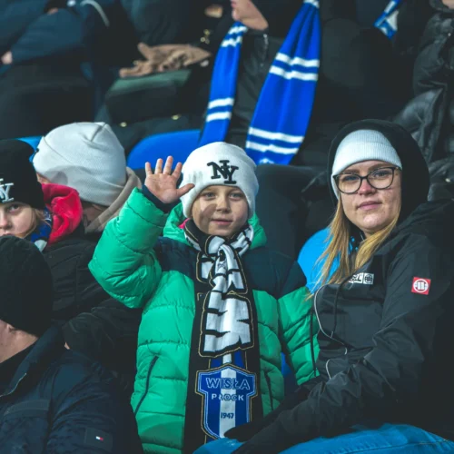 Na zdjęciu widoczny jest uśmiechnięty chłopiec w zielonym płaszczu trzymający szalik drużyny Wisła Płock, siedzący na trybunach z innymi kibicami.
