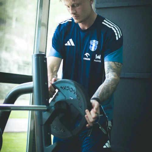 Mężczyzna w niebieskim stroju sportowym pracuje w siłowni, trzymając talerz ciężarowy przy sprzęcie do ćwiczeń.