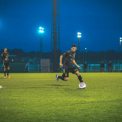 Na zdjęciu widoczna jest akcja na boisku piłkarskim, gdzie zawodnik w czarnym stroju biegnie z piłką, a jego rywale w pomarańczowych strojach próbują go dogonić.