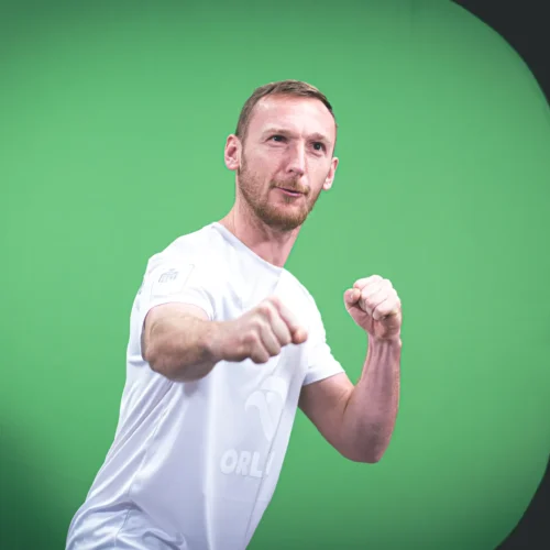 Mężczyzna w białej koszulce wykonuje pozę bojową na tle zielonej ściany.