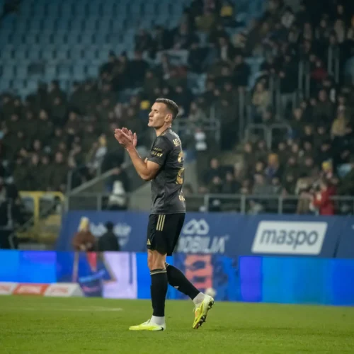 Na zdjęciu znajduje się piłkarz, który aplauzuje podczas meczu na stadionie, a w tle widać zgromadzoną publiczność.