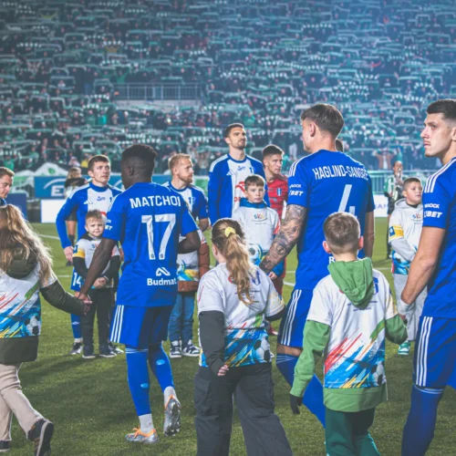 Na zdjęciu widać zawodników drużyny piłkarskiej w niebieskich strojach, którzy wychodzą na boisko, trzymając się za ręce z dziećmi.