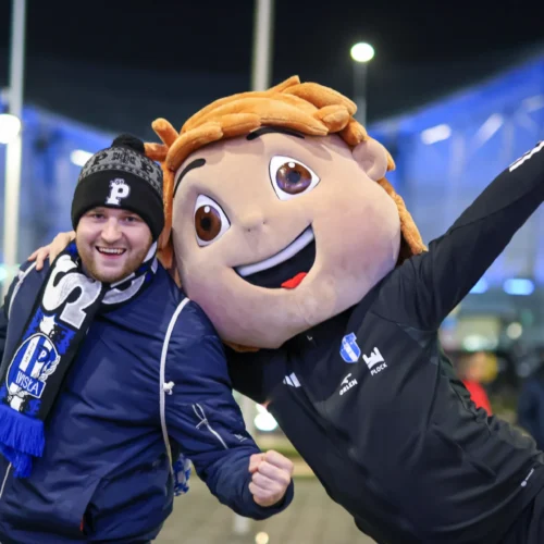 Na zdjęciu widoczny jest uśmiechnięty mężczyzna w czapce i szaliku, który pozuje z osobą w kostiumie maskotki, oboje wyrażają radość na tle nocnego otoczenia.