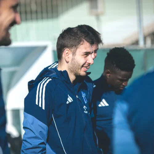 Na zdjęciu widać grupę mężczyzn ubranych w niebieskie stroje sportowe, którzy rozmawiają i się uśmiechają na boisku.