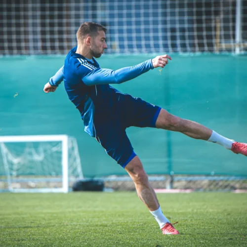 Mężczyzna w niebieskiej odzieży sportowej wykonuje kopnięcie piłki na boisku piłkarskim.