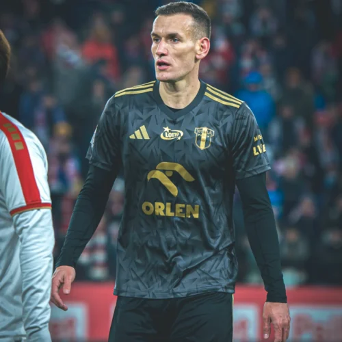 Na zdjęciu widoczny jest piłkarz w czarnej koszulce z złotymi elementami, stojący na boisku podczas meczu.