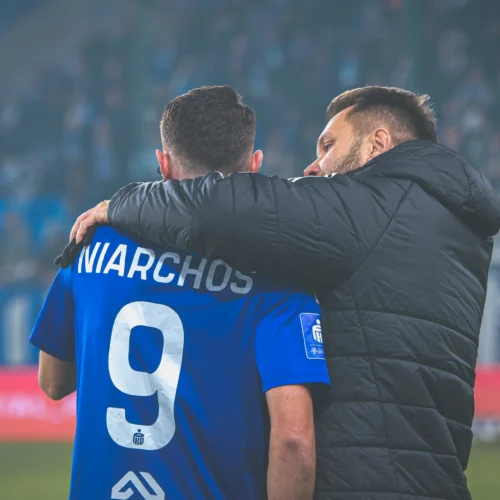 Na zdjęciu dwóch mężczyzn w niebieskich strojach sportowych w uścisku, jeden z nich ma na sobie numer 9, a w tle widoczna jest stadionowa atmosfera.