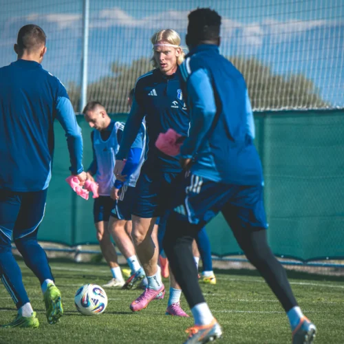 Na zdjęciu widać grupę piłkarzy ubierających się do treningu na boisku, z piłką oraz różowymi strojami treningowymi.
