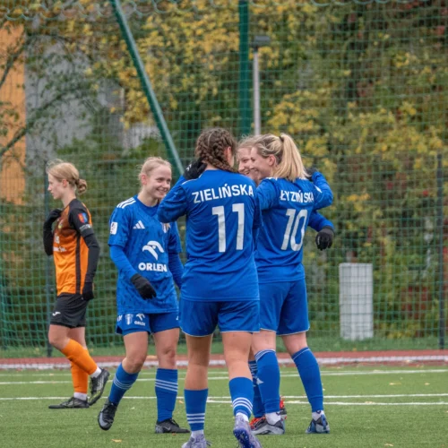 Na zdjęciu widoczna jest radosna grupa zawodniczek w niebieskich strojach piłkarskich, które świętują wspólne osiągnięcie na boisku.