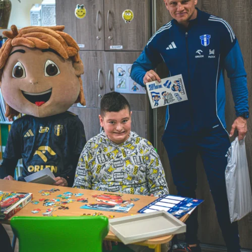 Na zdjęciu widoczny jest chłopiec siedzący przy stole z puzzlami, obok niego stoi osoba w kostiumie maskotki oraz mężczyzna w sportowym ubraniu, trzymający kartkę z naklejkami.