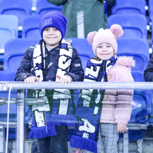 Dwoje dzieci w strojach sportowych z szalikami drużynowymi uśmiecha się do kamery, stojąc na trybunach.