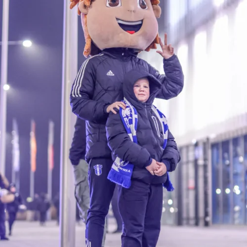 Na zdjęciu widać chłopca stojącego obok osoby w przebraniu postaci z dużą, uśmiechniętą głową oraz w niebiesko-białym szaliku, na tle nocnego otoczenia.