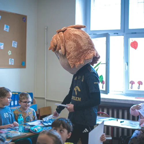 Na zdjęciu w klasie widać osobę w przebraniu z dużą głową, która rozmawia z uczniami siedzącymi przy biurkach.
