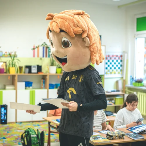 W klasie szkolnej stoi postać w dużej masce z włosami, trzymająca kartki, podczas gdy dzieci w tle są zaabsorbowane swoimi zajęciami.