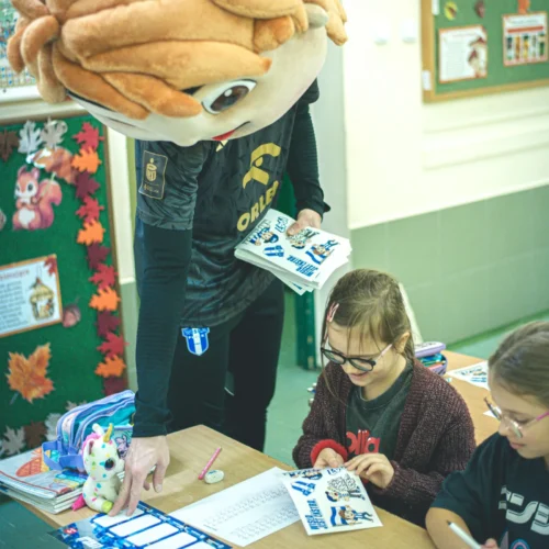 Na zdjęciu widoczny jest nauczyciel w przebraniu maskotki, który przekazuje kolorowanki dwóm uśmiechniętym dziewczynkom w klasie szkolnej.