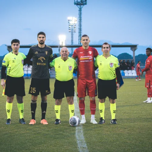 Na zdjęciu widać dwóch piłkarzy, sędziów oraz asystenta sędziego przed meczem piłkarskim na boisku, z górzystym tłem.