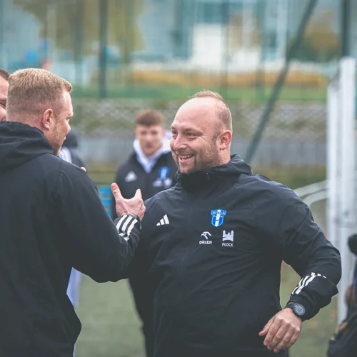 Na zdjęciu widać grupę mężczyzn w czarnych kurtkach, rozmawiających i uśmiechających się na boisku piłkarskim.