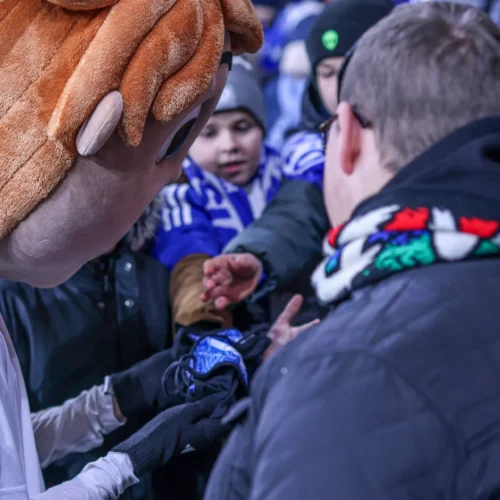 Na zdjęciu widoczny jest człowiek w kostiumie maskotki, który rozdaje pamiątki grupie dzieci i dorosłych na stadionie.