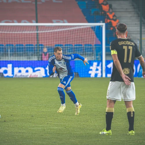 Na zdjęciu widać dwóch piłkarzy podczas meczu, z jednym z zawodników w niebieskiej koszulce biegnącym w kierunku piłki.