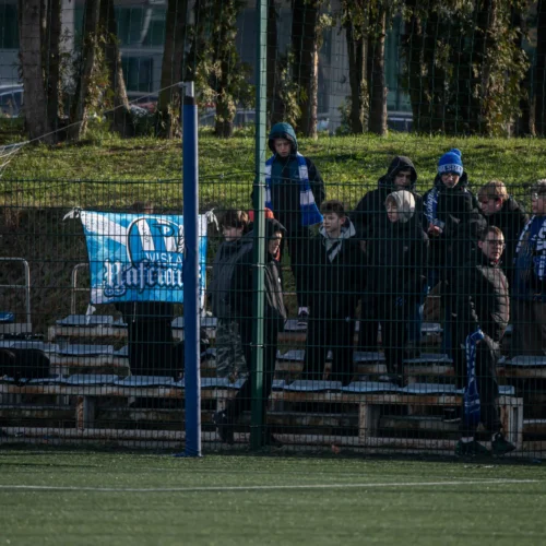 Na zdjęciu widać grupę kibiców stojących na trybunach, z flagą i transparentem klubu sportowego w tle.