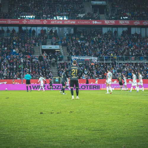 Mecz piłkarski z dużą ilością kibiców na trybunach, z widocznymi zawodnikami obu drużyn na boisku.