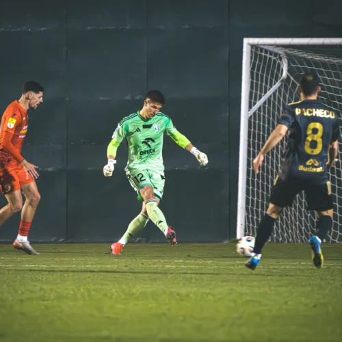 Na zdjęciu widać dwóch piłkarzy na boisku, z których jeden jest bramkarzem w zielonym stroju, a drugi w pomarańczowym, podczas intensywnej akcji meczowej.