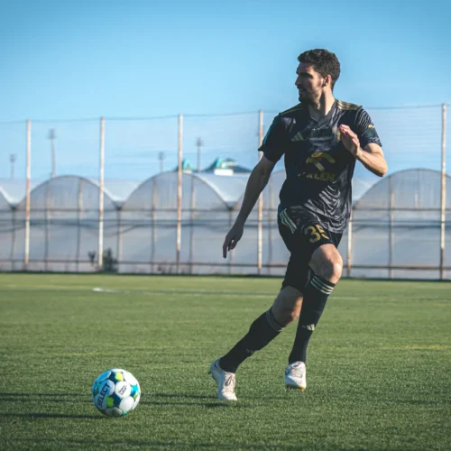 Na zdjęciu widać piłkarza w czarnej koszulce, który biegnie w kierunku piłki na zielonym boisku.