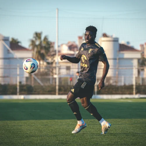 Na zdjęciu widoczny jest piłkarz w czarnej koszulce treningowej, który gra z piłką na boisku.